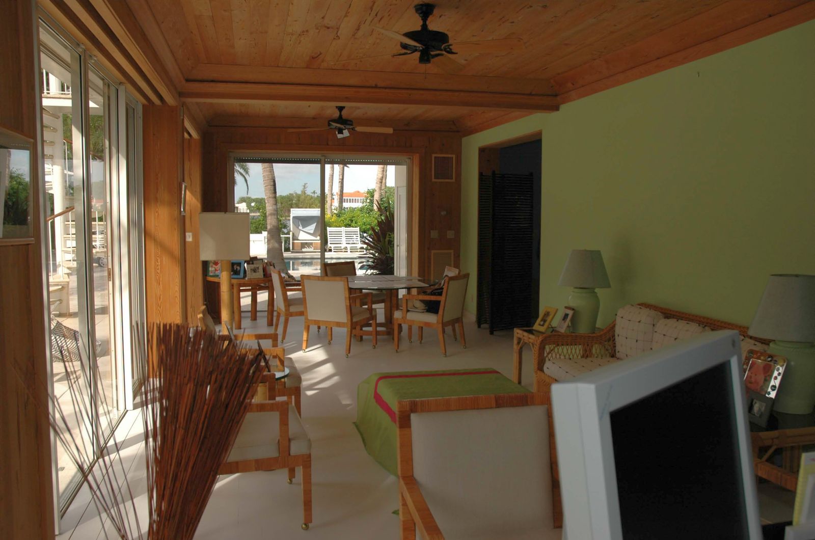 A wood-ceilinged living space with wicker furniture opening through sliding glass doors onto palm trees and a pool