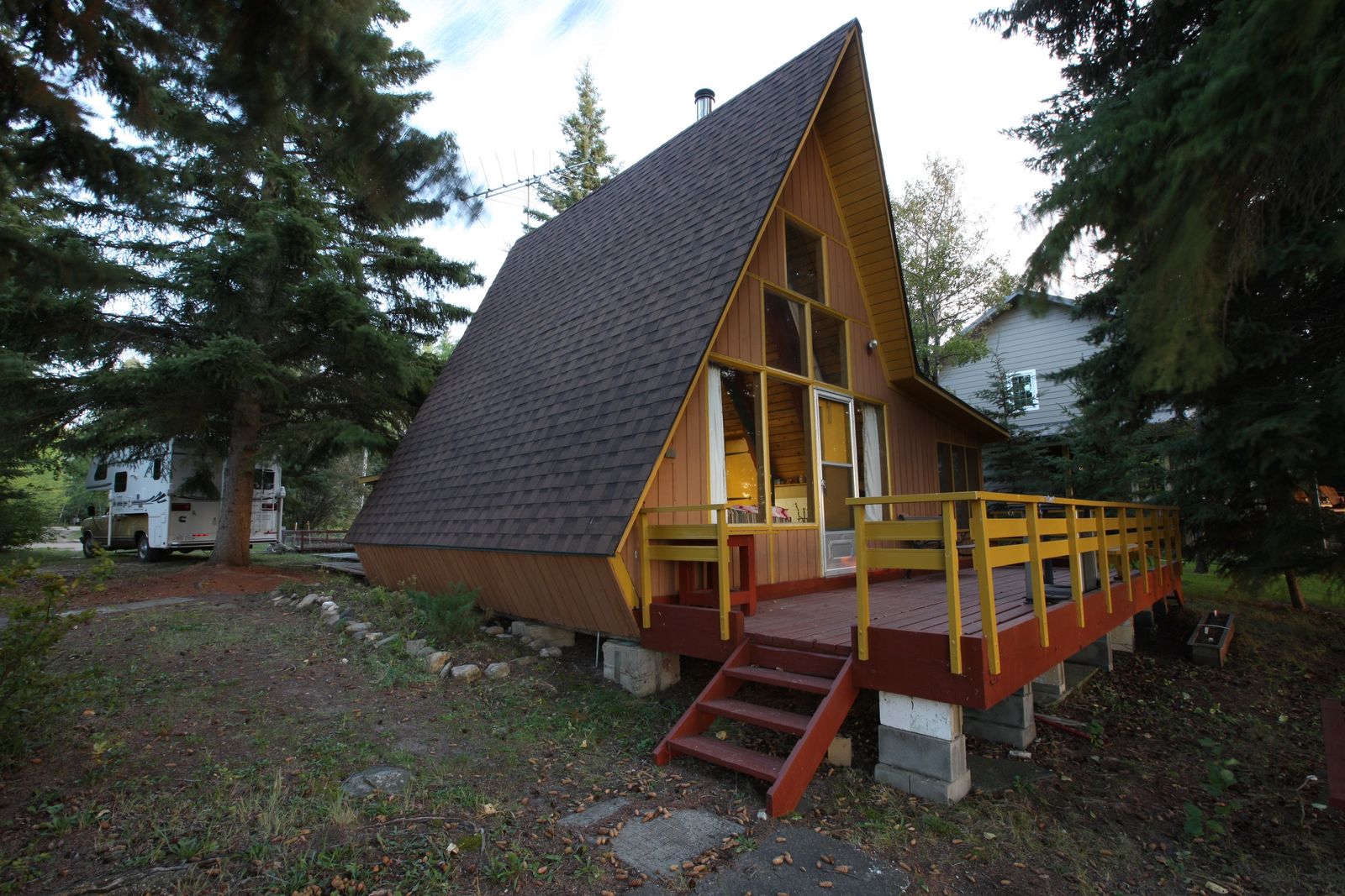 A classic yellow-trimmed A-frame cabin among pine trees, raised on concrete-block piers with a wooden deck and stairs