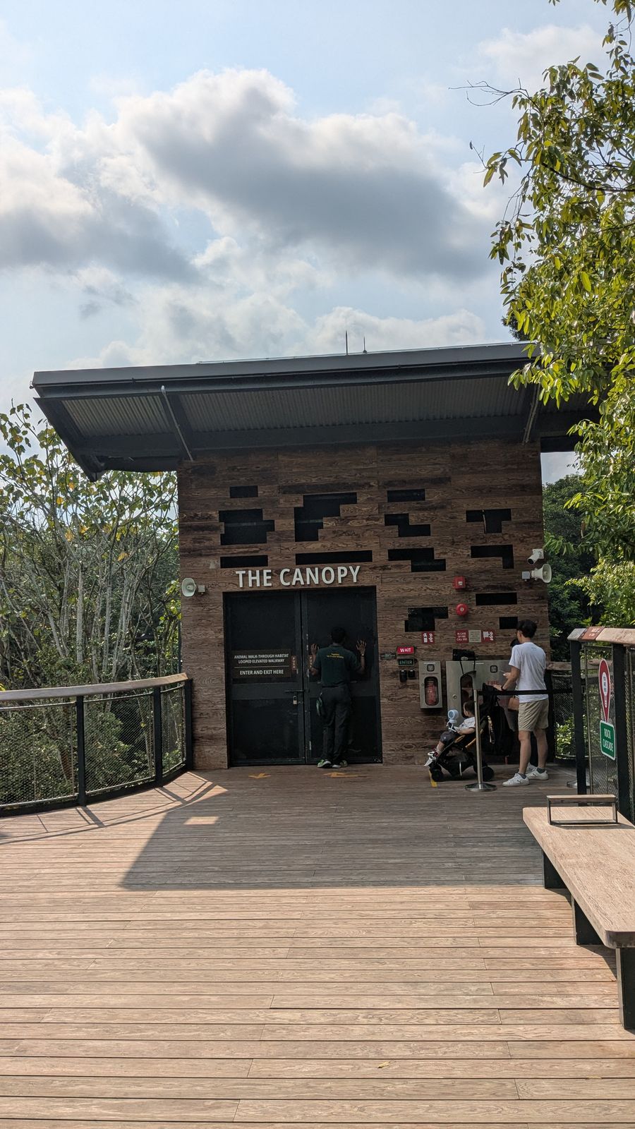 A wood-clad building with a sign reading 'The Canopy' at the entrance to an elevated wooden walkway, with visitors gathered at the doorway