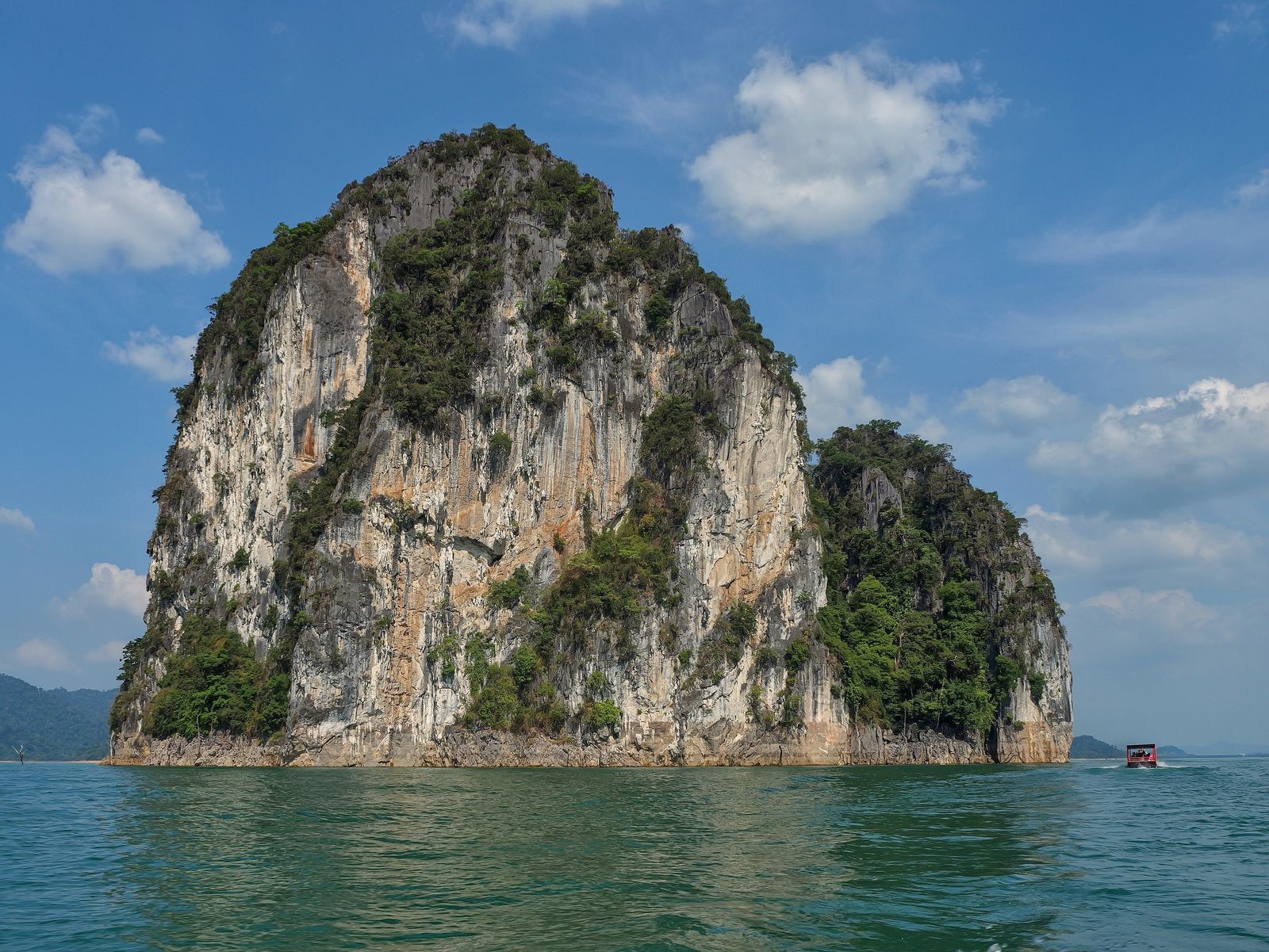 A limestone karst island rising from the turquoise water of Cheow Lan Lake in Khao Sok National Park, Thailand, with a longtail boat passing at its base
