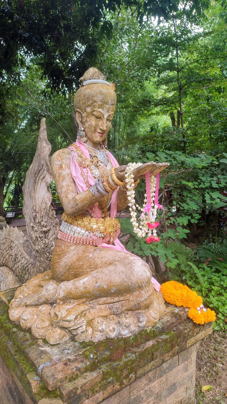 A gilded Thai deity statue set among dense jungle greenery at a forest temple, adorned with garlands
