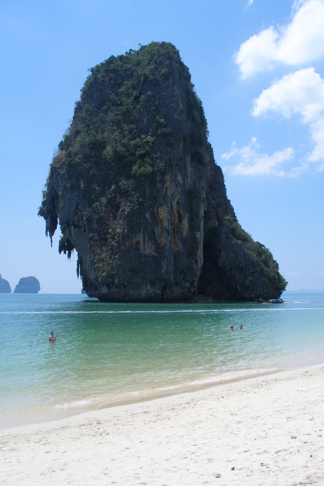 A tall limestone karst tower draped in jungle vegetation rising from the sea at Railay, Krabi, Thailand, with swimmers and a longtail boat in the foreground