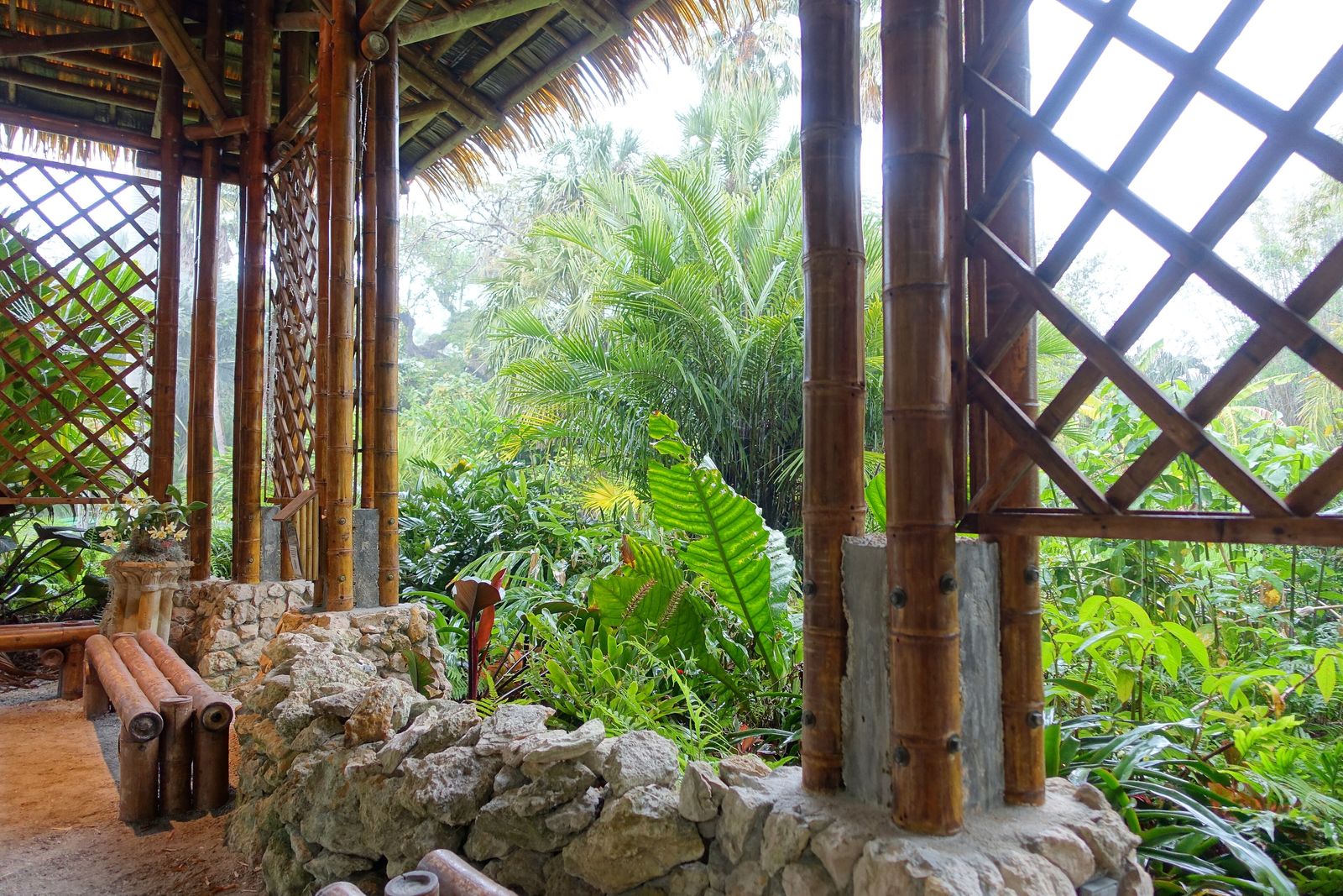 An open bamboo pavilion with lattice screen walls and a thatched roof, its posts anchored on stone footings above dense tropical planting